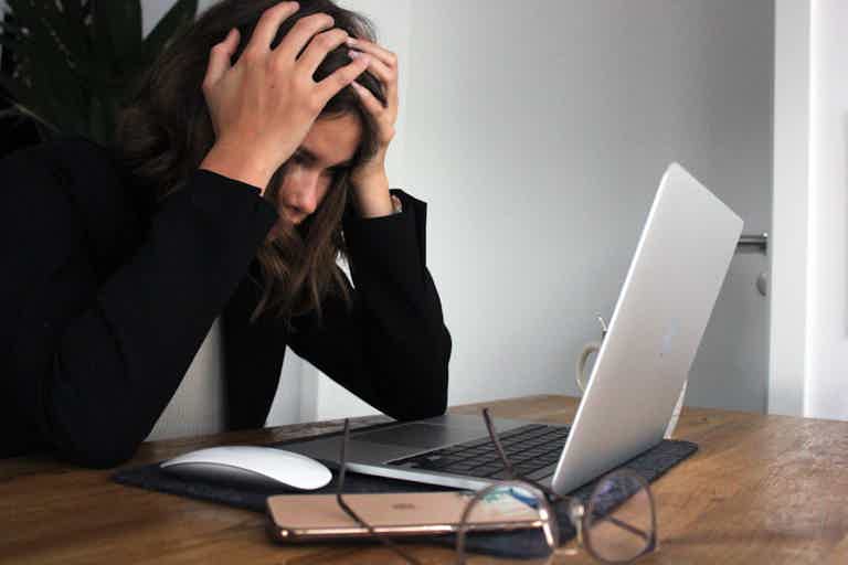 Woman sitting at desk, hands on head in distress, looking at an open laptop with a smartphone and glasses nearby.