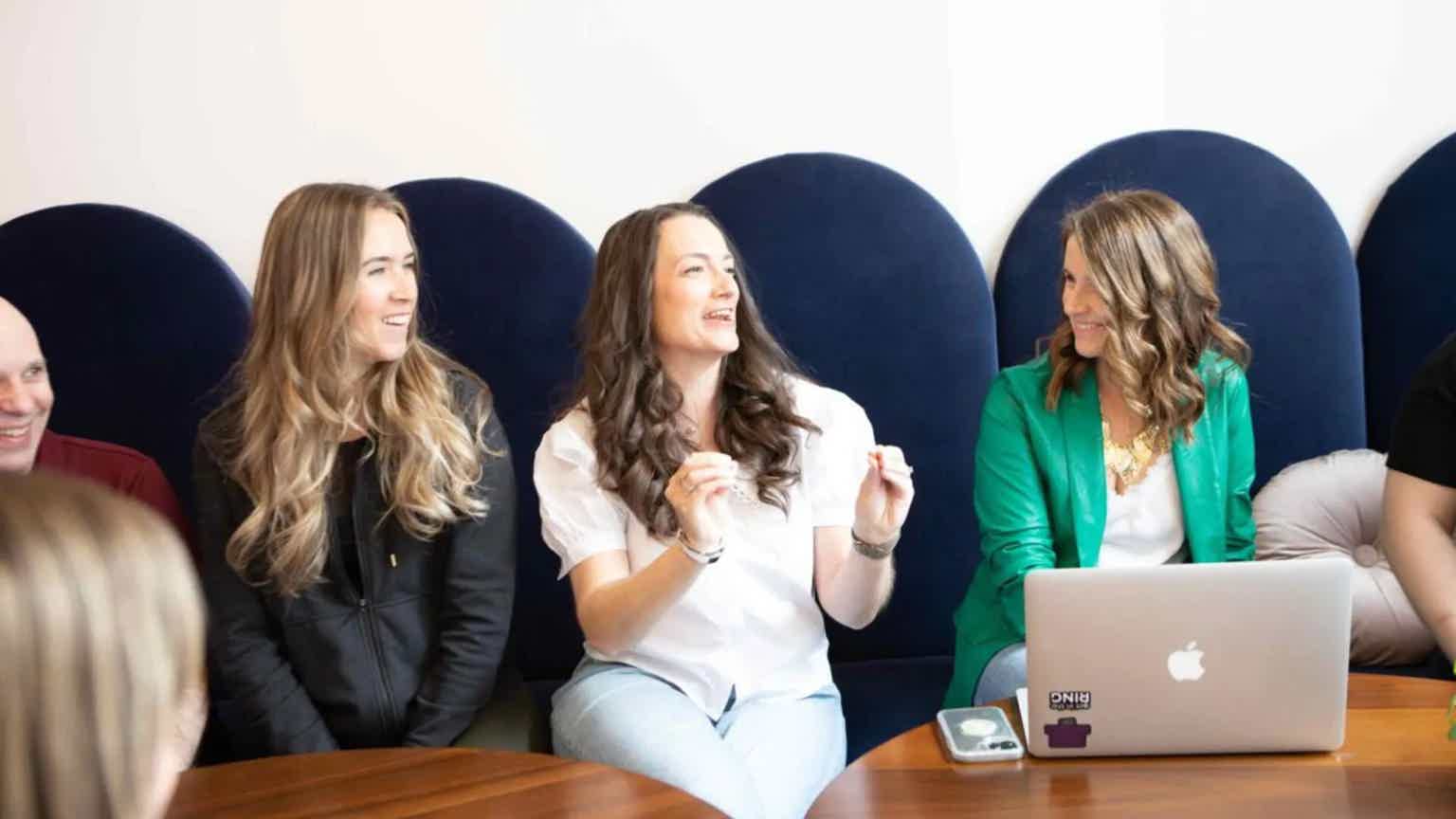 Four people seated in blue theater seats, smiling and laughing during a meeting with a laptop on a table.