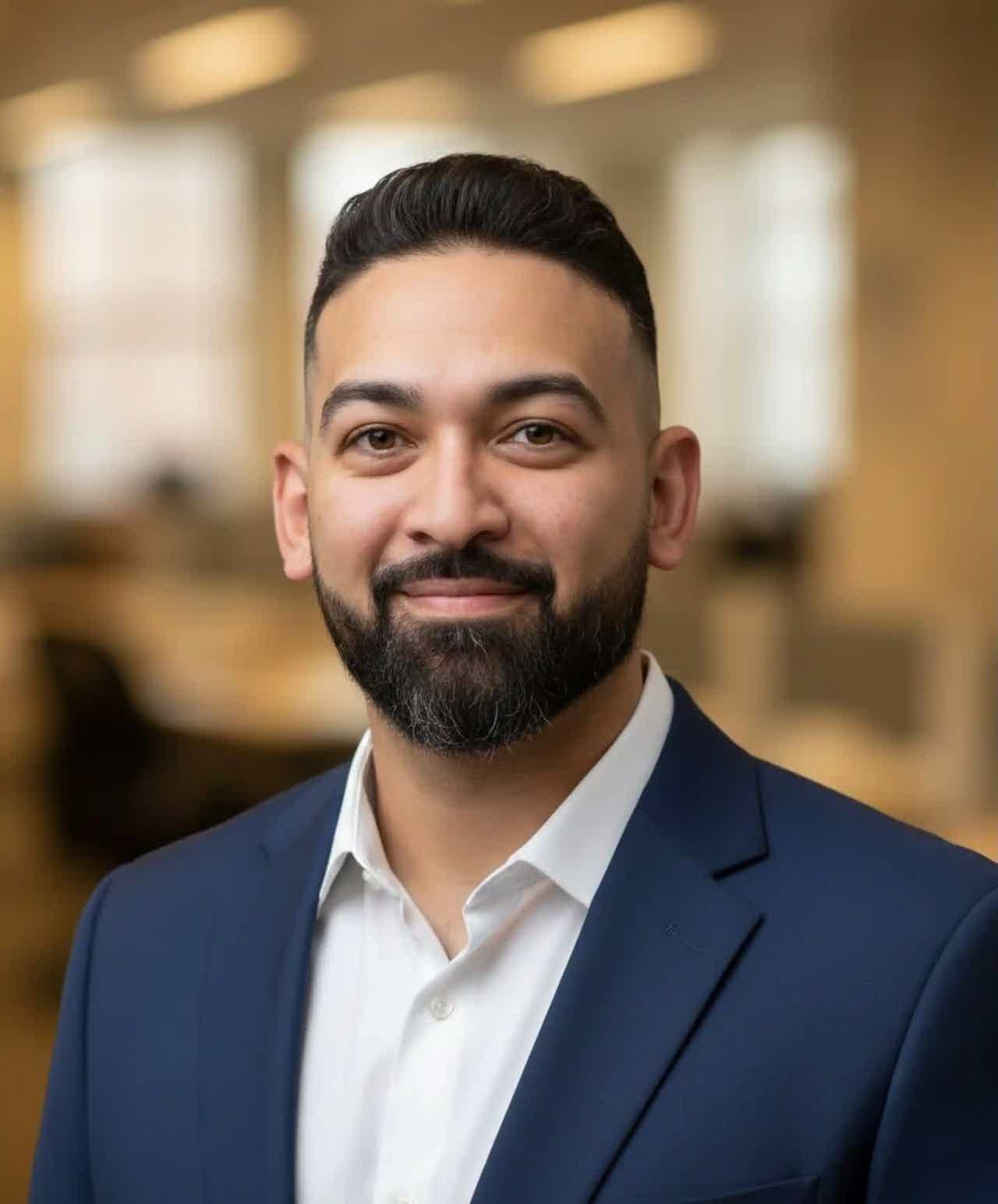 Smiling man with dark hair and full beard wearing a dark blazer and white shirt against a light background.