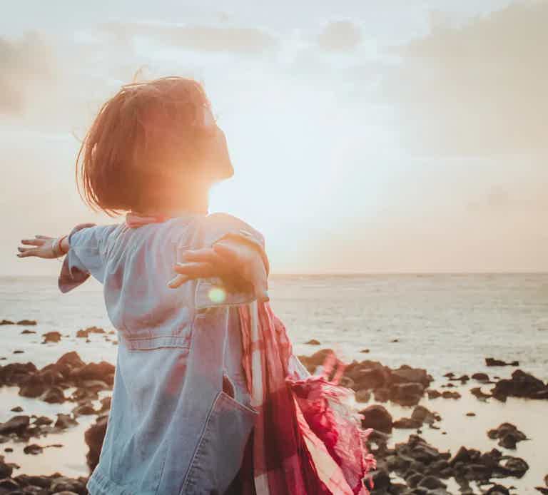 Child at the beach with arms outstretched, facing sunlit horizon, wearing a light denim dress and a pink scarf.