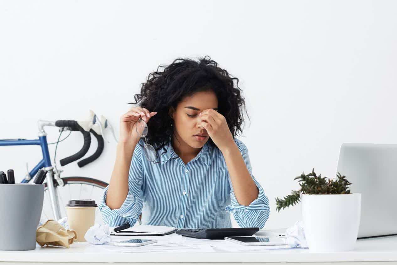Fatigued woman rubbing her eyes at a cluttered desk surrounded by crumpled papers and a laptop.
