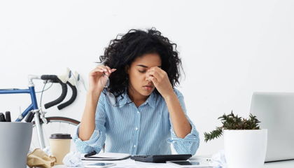 Fatigued woman rubbing her eyes at a cluttered desk surrounded by crumpled papers and a laptop.