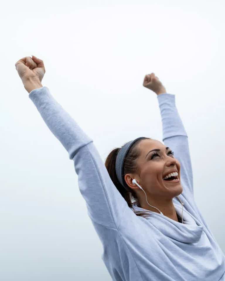 Athletic woman in light hoodie, headband, and earphones, raising arms in celebration against a pale sky.