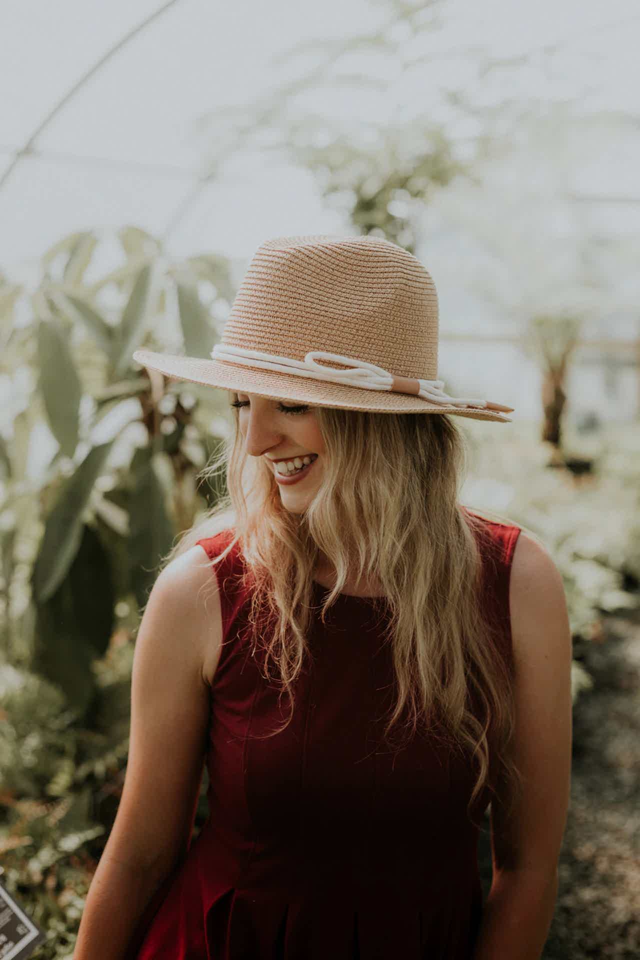 Woman in a straw hat and burgundy dress smiles in a greenhouse with plants around.